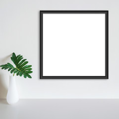 Black frame mockup on white wall with philodendron leaf in vase on white surface. Copy space.