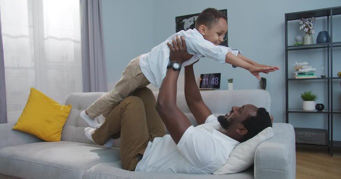 Joyful man lifting his small cute son up like flying at home indoor. Handsome happy African American young father playing plane game with little boy while lying on couch in room. Family concept