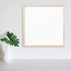 Wood frame mockup on white wall with philodendron leaf in vase on white surface. Light wood. Copy space.