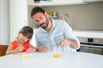 Young dad holding brush with fingers and playing with daughter in kitchen. Cute girl painting Easter egg, sitting and drawing with Latin father. Family time, hobby, childhood and holiday concept