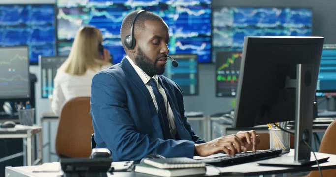 Handsome African American man in headset. Broker working at monitor of computer and speaking with client. Male stock trader talking on phone in trading office and typing on keyboard. Sales department.
