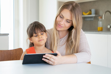 Fototapeta na wymiar Beautiful mom and her daughter watching movie via tablet together. Adorable little girl sitting at kitchen with young pretty mother and looking at screen. Family and digital technology concept