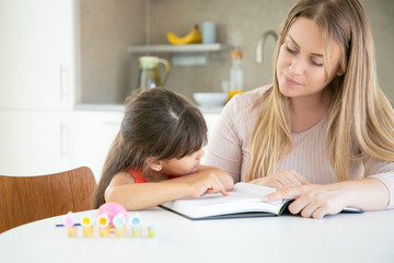 Obraz na płótnie Canvas Lovely cute girl pointing at text and asking mom about something. Beautiful young mother teaching little daughter and reading book with her in kitchen. Family time, childhood and education concept