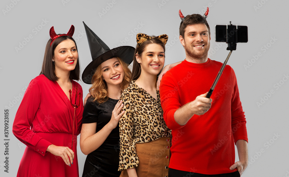 Poster friendship, holiday and people concept - group of happy smiling friends in halloween costumes of witch, devil and cheetah taking picture by smartphone on selfie stick over grey background