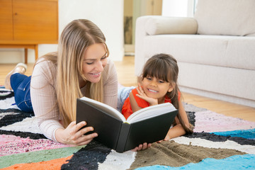 Pretty mother lying on rug with daughter and reading book to her. Adorable little girl smiling, learning to read with mom, watching on text and listening story. Family time and education concept