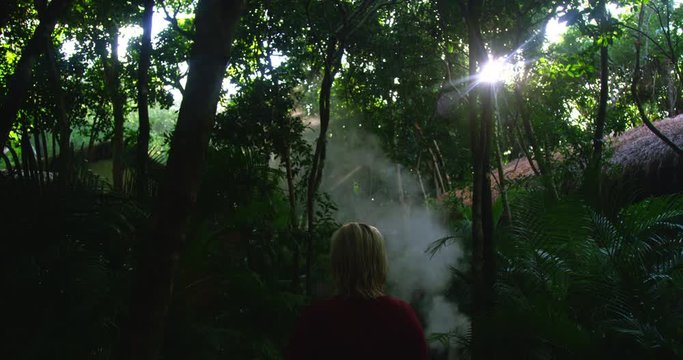 Sunlight glistens shining through palm trees revealing woman standing at the edge of a jungle forest path surrounded by rich green leaves and trees