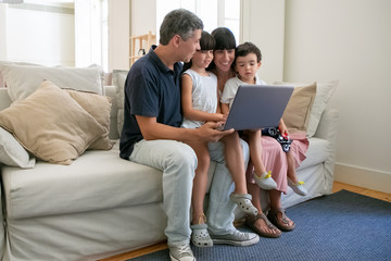 Happy parents couple holding kids on lap, sitting on couch all together, watching movie or video on laptop at home. Full length. Communication or entertainment concept