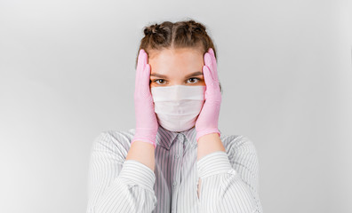 Beautiful young female in a white shirt, in a medical mask and pink sterile gloves is holding head in hands. She shocked and holds her head. The concept of self-isolation, quarantine and pandemic