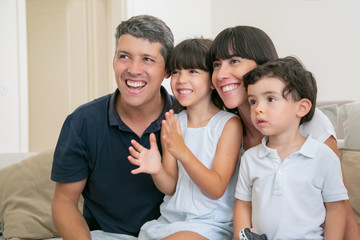 Excited joyful parent couple with two kids watching TV, sitting on couch in living room, looking away and smiling. Medium shot. Leisure time with family or television concept