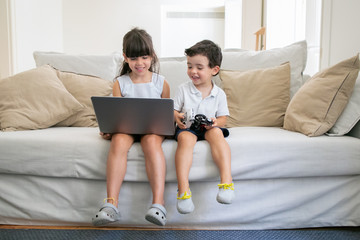 Happy joyful boy and girl sitting on couch at home, using laptop, watching video, cartoon films or...