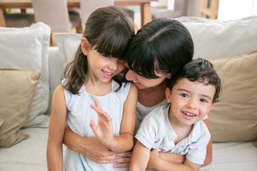 Obraz premium Happy mom enjoying time with kids at home. Cheerful mother and two children sitting on couch in living room together. Leisure time with family concept