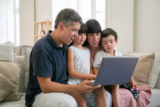 Excited cheerful parents couple holding kids on lap, sitting on couch all together, watching movie or video on laptop at home, staring at display. Medium shot. Communication or entertainment concept