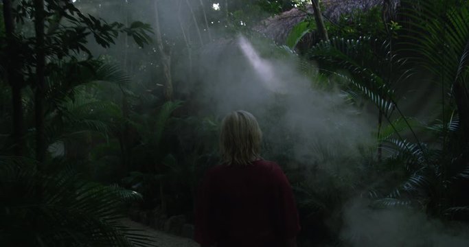 Mysterious woman walks along dense forest path surrounded by dark green leaves and trees through fog and mist