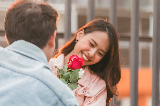 Young Happy Couple Love And Romantic At First Date Relationship. Asian Teenage Woman Surprise And Smiling At Boyfriend Gives Red Rose Flowers At Dinner In Valentine Day. Focus On Rose Flower.