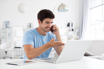Happy smiling man works on his laptop and talking phone at home. Cheerful freelance.