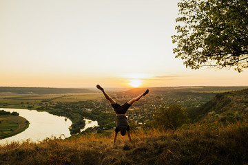 Man doing handstand on the grass at sunset sky. Young sporty man doing handstand exercise in beautiful mountain landscape. Sports outdoor lifestyle.  Copy space.