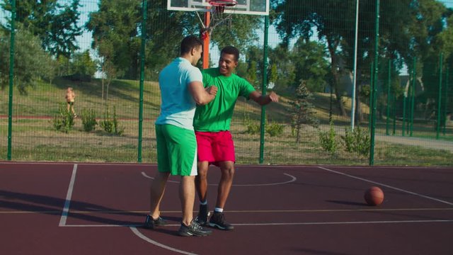 Sporty fit mixed race streetball player extending hand to lift fallen opponent player off ground, helping to stand up , practicing good sportsmanship during basketball game on outdoor court.