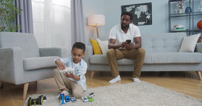 Concentrated African American handome man typing and browsing on smartphone while cute boy playing with toys on floor in cozy room at home. Little child having fun. Man tapping on cellphone