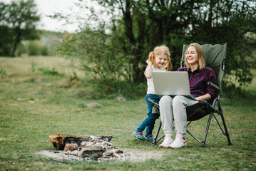Mother work on Internet with kid outdoors. Quarantine, closed nursery school during coronavirus outbreak. Communication with family online on laptop near fire in nature. Homeschooling, freelance job.