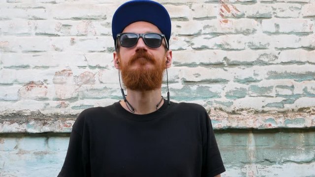 Portrait of young red bearded man in black t-shirt and snapback in rock style on urban background listening to music with headphones. Hipster guy in wireless earphones on old blue brick wall backdrop.