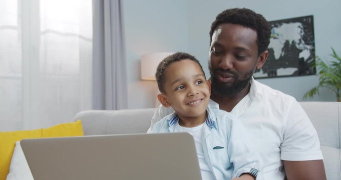 Close up portrait of handsome joyful male typing on laptop while sitting on sofa with small son in room. African American happy father with cute boy tapping on computer while sitting at home indoors