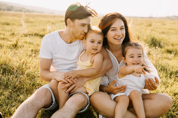 Mother, father and two daughters sitting outdoors in an open field with green grass, smiling.