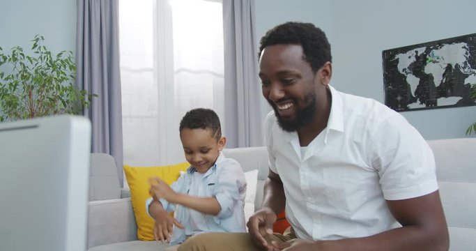 Portrait of happy African American handsome man playing with little cute son with ball while sitting on sofa in room. Cheerful boy spends time with his dad at home. Happy family. Childhood concept