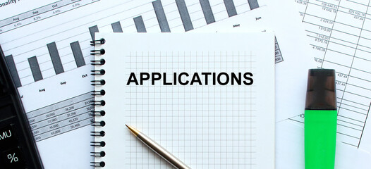 Text APPLICATIONS on the page of a notepad lying on financial charts on the office desk. Near the calculator and marker. Business concept.