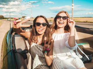 Portrait of two young beautiful and smiling hipster girls in convertible car. Sexy carefree women driving cabriolet. Positive models riding and having fun in sunglasses