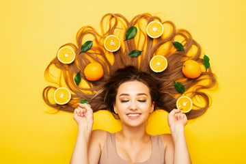Obraz na płótnie Canvas happy girl lying on colored background with orange fruits on long hair, young surprised woman with citrus slices and leaves, concept good news