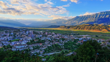 Fototapeta na wymiar View of Gjirocaster city in shadow from Gjirocaster castle with green fields and monumental mountain ridge in the background during beautiful sunset, Gjirocaster, Albania
