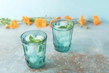 Two beautiful vintage turquoise glasses with cold drink and ice cubes, with fresh green mint leaves decorated with marigold flowers on natural colorful stone table. Summer concept.