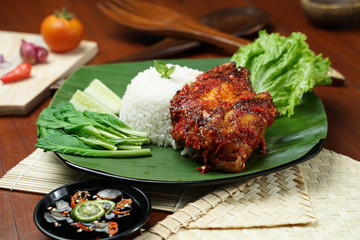 Tasty spicy grilled juicy chicken with golden red skin served with white rice, chili sauce and various vegetables. with a beautiful bamboo plate on a wooden table.