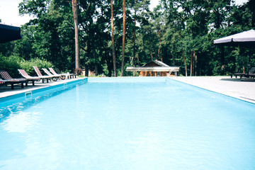 View of the water surface in the pool from the side against the background of green trees. Surface of blue swimming pool. Background of blue ripped water in swimming pool