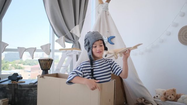 Little boy in pilot hat playing with model of plane sitting in box indoors
