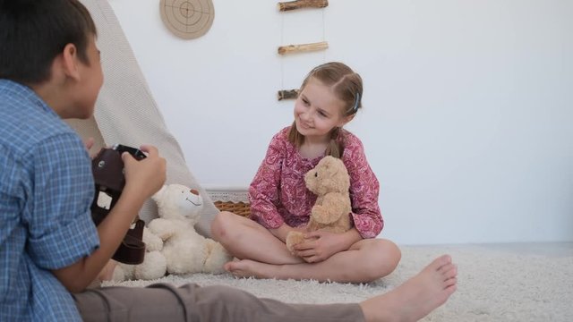 Little boy photographing little girl with retro camera sitting on floor in light room with teddy bear