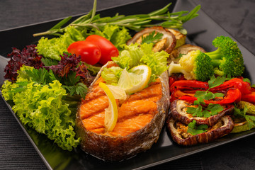 Serve food in the restaurant. Food poster - large portion of red fish, trout, salmon steak with grilled vegetables, salad, broccoli and tomatoes, decorate with lemon and rosemary.