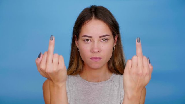 Serious woman in gray t-shirt shows middle finger on blue background with copy space. 4K slow motion footage