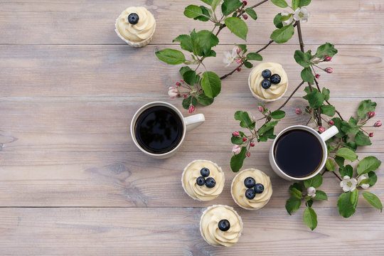 Two beautiful porcelain coffee cups with homemade blueberry cupcakes with vanilla cream on wooden table in spring garden. Apple tree branches in bloom in setting sun light. Flat lay style.