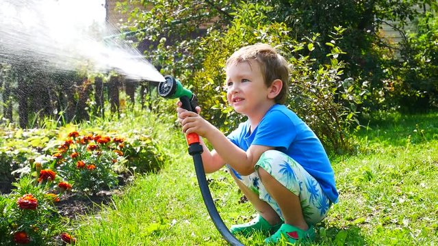 A little boy waters the garden flowers with a watering spray sitting on the lawn