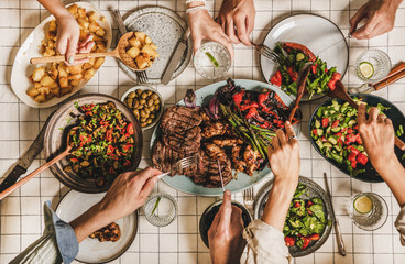 Summer barbeque party. Flat-lay of table with grilled meat, vegetables, salad, roasted potato and peoples hands with cutlery over white tablecloth, top view. Family gathering, comfort food concept - obrazy, fototapety, plakaty