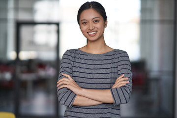 Fototapeta premium Portrait of a young happy asian woman, female office worker in casual clothes keeping arms crossed, looking at camera and smiling while standing in the modern office
