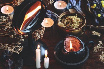 Obraz na płótnie Canvas Candle flame divination in concave mirror on wiccan witch altar. Black candle burning on a messy cluttered table with dried herbs and flowers, crystals, plants, other nature objects in dark background