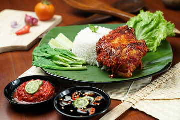 Tasty grilled juicy chicken with golden brown skin served with plain white rice, chili sauce and various vegetables. with a beautiful bamboo plate on a wooden table.
