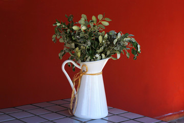 White enamel vase with leaf branches on ceramic tile table. Orange colored wall in the background.