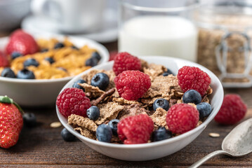 Corn flakes with blueberries and raspberries in white bowl in dark wooden desk.