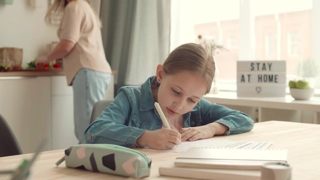 Medium shot of blond-haired Caucasian schoolgirl sitting at kitchen table and doing homework while her mother cooking on background. Then woman coming and starting helping her daughter