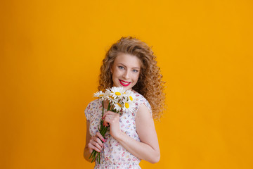 a curly blonde woman holding a bouquet of daisies on a yellow background smiles