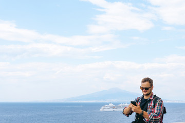Male tourist with glasses and a banana bag, uses a smartphone. Communication and mobile internet concept. Roaming. View of the sea and Mount Vesuvius. Ocean liner.