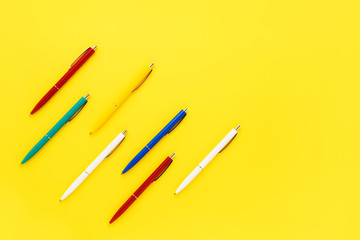 Seven colorful ballpoint pens on a bright yellow background. Top view, copy space for text.